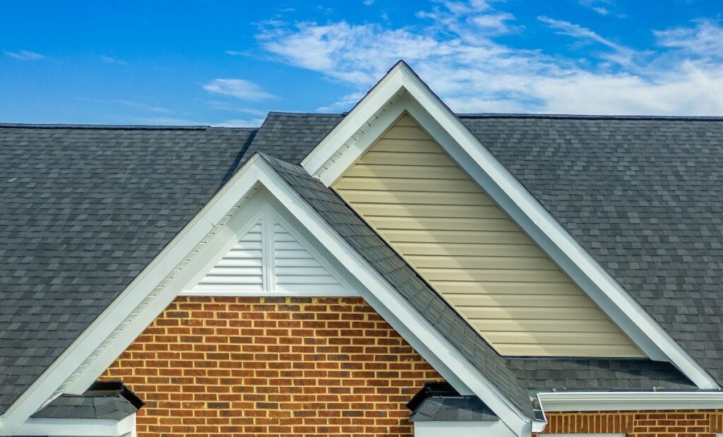Double gable with beige horizontal vinyl lap and red brick facade siding, with triangle shape white attic vent on a pitched roof attic at an American single family home neighborhood USA