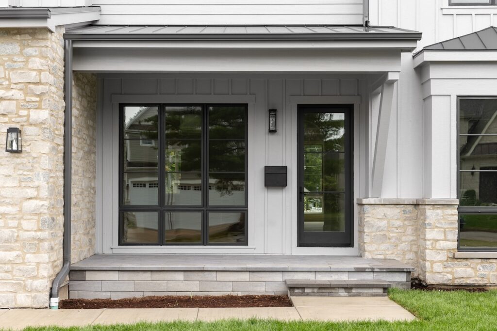 A modern farmhouse front door detail with large black framed windows and front door, grey board and batten siding, and stone front porch and siding.