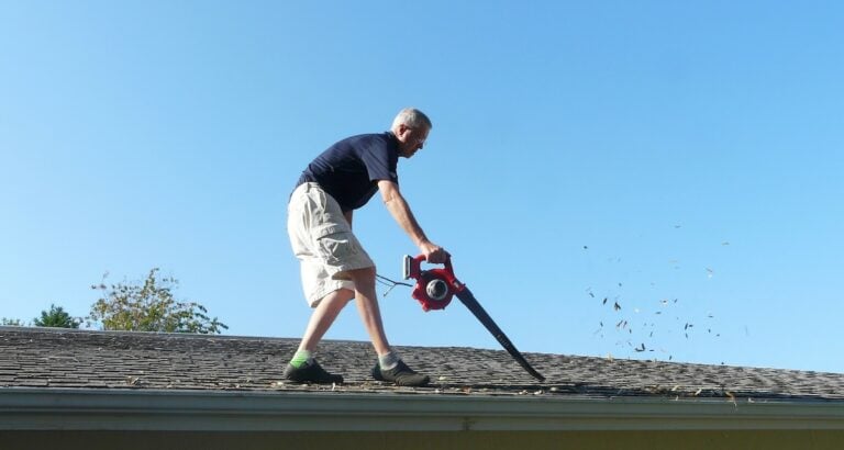 A homeowner is on the roof of his residence using a leaf blower to remove leaves from the gutter in preparation for storm season water runoff.