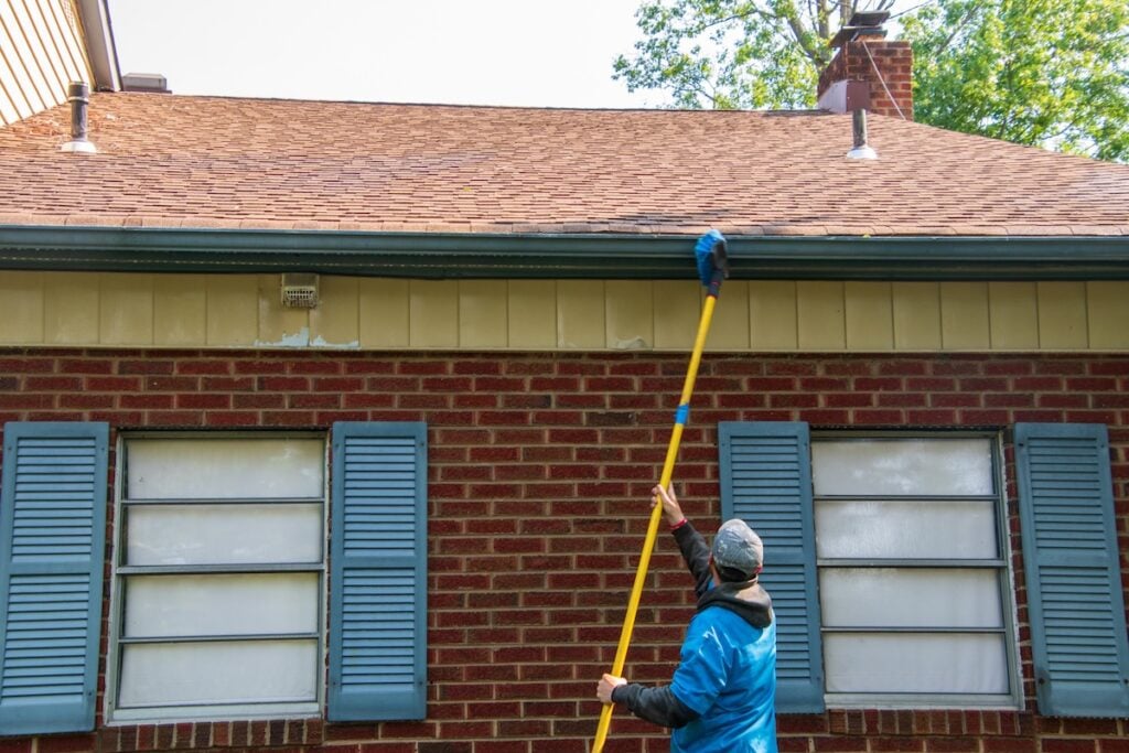 Young man cleaning the soffit of a house with a brush on a long pole. The house has blue shutters and is one story high