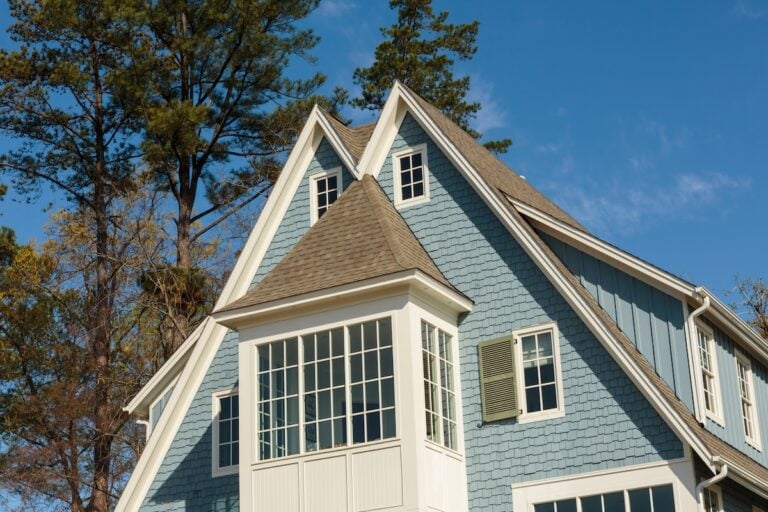 Upward view of blue home's dual peaked roof of attic, with trees in the background. House is blue with shingles, with white detail.
