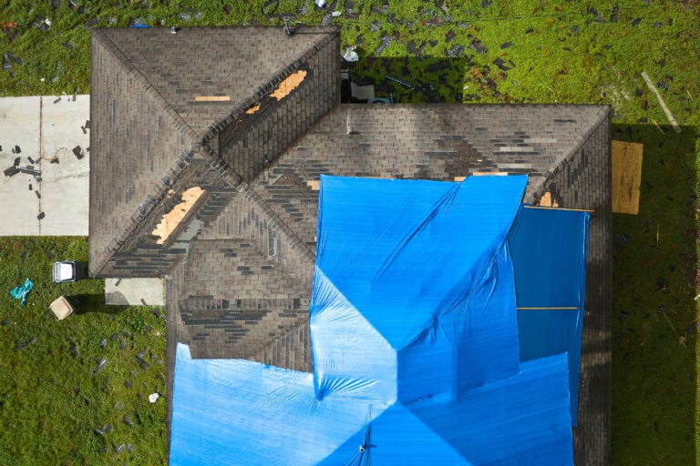 Aerial view of damaged in hurricane Ian house roof covered with blue protective tarp against rain water leaking until replacement of asphalt shingles.